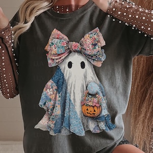 May include: Dark grey t-shirt with a ghost design. The ghost has a floral bow and patchwork clothing, holding a pumpkin basket. The design has a vintage, slightly distressed look, ideal for Halloween.