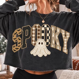 May include: A charcoal gray long-sleeve cropped sweatshirt featuring the word SPOOKY in large block letters. The letters have varied patterns including bats, black glitter, brown plaid, black and white checks, and small white ghosts.