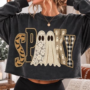 Op de afbeelding: Een antracietgrijze cropped sweater met lange mouwen en het woord SPOOKY in grote applicatieletters. De S heeft een vleermuispatroon, de P is zwart, de twee O's zijn spookvormen met kant- en stoftexturen, de K heeft kleine spookjes en de Y is geruit.