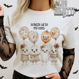 Puede incluir: Camiseta blanca con la frase "Hangin' With My Boos" y un gráfico de cuatro personajes fantasma sosteniendo globos. Los globos y los disfraces de fantasma tienen diseños florales y de calabaza, ideal para Halloween.