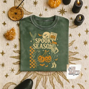 May include: Green crew neck sweatshirt with a Halloween-themed graphic. The design includes the words "Spooky Season", a ghost, pumpkins, bats, and a cowgirl. The sweatshirt has a vintage, washed look.