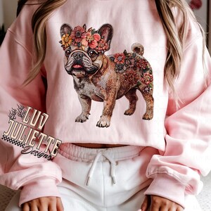 Puede incluir: Una sudadera rosa con un dise&ntilde;o floral de un bulldog franc&eacute;s. El bulldog lleva una corona de flores. La sudadera tiene el texto "Luv Juliette Designs" en la esquina inferior izquierda.