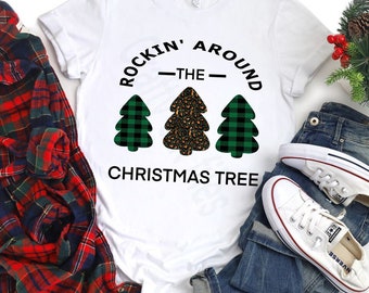 May include: White t-shirt with the text "ROCKIN' AROUND THE CHRISTMAS TREE". The design features three Christmas trees: two green plaid trees and one leopard print tree. The shirt is paired with jeans, sneakers, and a plaid shirt.