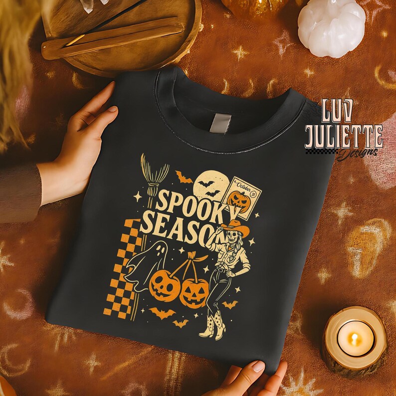 May include: Black crewneck sweatshirt with a Halloween-themed graphic. The design features the text "Spooky Season" with a ghost, pumpkins, bats, and a cowgirl. The sweatshirt is held by a person.