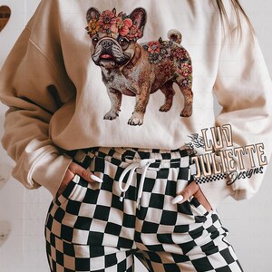Puede incluir: Una sudadera beige con un dise&ntilde;o floral de un bulldog franc&eacute;s con una corona de flores. La sudadera la lleva puesta una persona que lleva pantalones de cuadros negros y blancos.