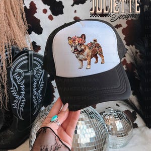 Puede incluir: Una gorra de camionero blanca y negra con un dise&ntilde;o floral en la parte delantera. El dise&ntilde;o presenta un bulldog franc&eacute;s con flores en la cabeza. El texto "LUV JULIETTE Design" es visible en la gorra.