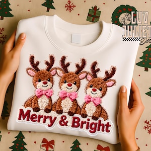 May include: A white sweatshirt featuring an embroidered design of three reindeer with brown antlers and pink bow ties. The text "Merry & Bright" is embroidered below the reindeer. The sweatshirt is held up by two hands.