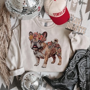 Puede incluir: Una sudadera beige con un dise&ntilde;o de bulldog franc&eacute;s con estampado floral. La sudadera est&aacute; sobre una alfombra estampada con una bola de discoteca y botas de vaquero.