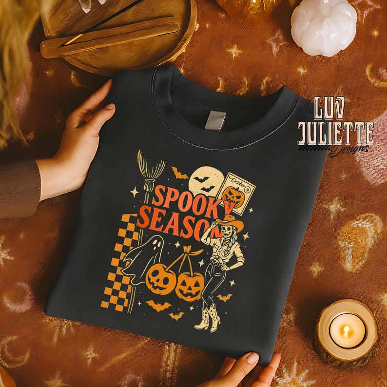 May include: Black crewneck sweatshirt with a Halloween-themed graphic. The design features the text "Spooky Season" with a ghost, pumpkins, bats, and a skeleton cowgirl. The sweatshirt is held by a person.