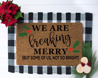 May include: A rectangular doormat with the text "WE ARE freaking MERRY (BUT SOME OF US, NOT SO BRIGHT)" in black script. The mat is brown with green holly and red berries accents. It sits on a black and white checkered rug, with red poinsettias and pine sprigs.