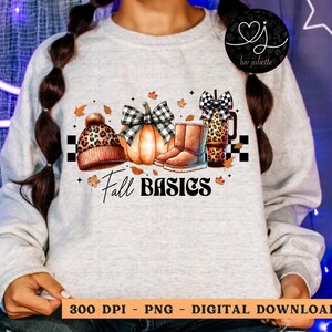 May include: A white sweatshirt with a graphic design of fall-themed items, including a pumpkin, boots, a hat, and a mug. The text "Fall Basics" is printed below the graphic.