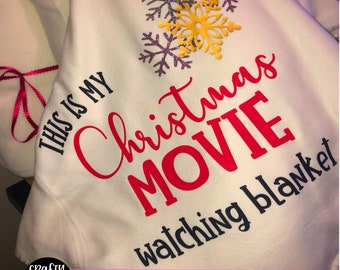 May include: White fleece blanket with black text that reads "This is my Christmas Movie watching blanket". The text is surrounded by three glittery snowflakes in silver and gold.