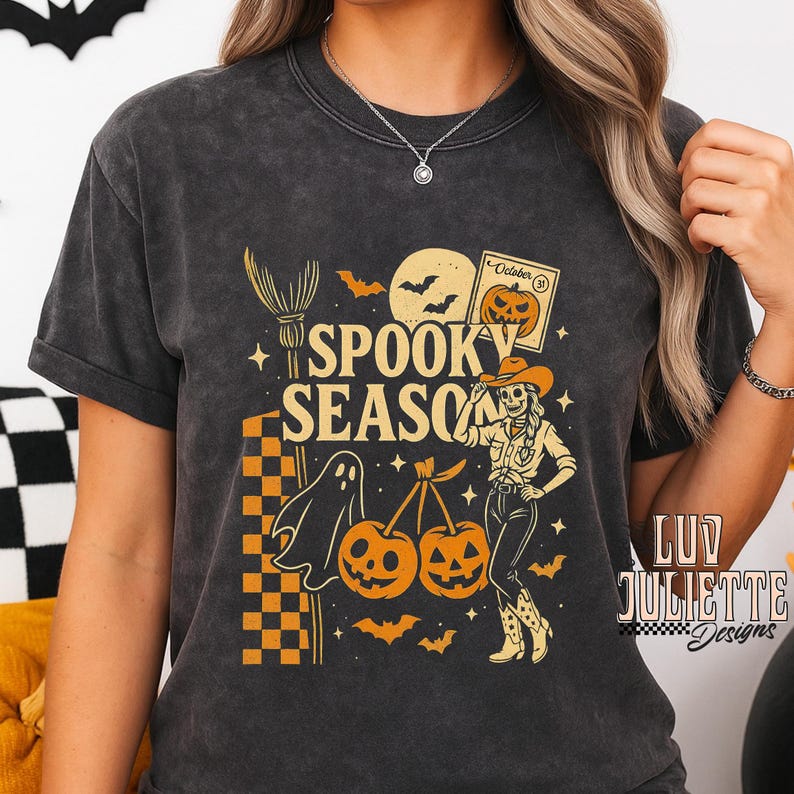 May include: Dark gray t-shirt with a Halloween-themed graphic. The design includes the text "Spooky Season", a ghost, pumpkins, bats, and a skeleton cowgirl. The shirt also features a broom, a full moon, and a calendar.
