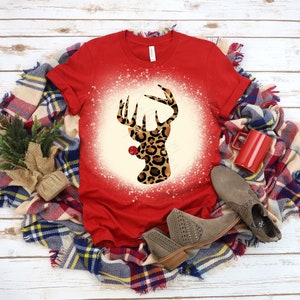 May include: A red t-shirt featuring a deer head design with a leopard print pattern. The deer head is centered on a cream-colored circle with a red border. The shirt is displayed on a plaid blanket with a red mug and gray boots.
