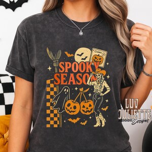 May include: Dark gray t-shirt with a Halloween-themed graphic. The design features the text "Spooky Season" with a witch's broom, a ghost, pumpkins, bats, and a skeleton cowgirl. The design includes orange, yellow, and black colors.