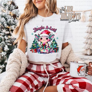 Könnte beinhalten: Weißes T-Shirt mit einer farbenfrohen Grafik eines festlichen Axolotls mit Weihnachtsmütze, das ein Geschenk hält, und dem Text "Jingle A-Lot!". Der Axolotl wird von Weihnachtsbäumen und Geschenken flankiert. Die Person trägt eine rot-weiß karierte Pyjamahose.