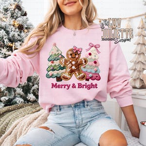 May include: A light pink sweatshirt featuring a gingerbread man and two Christmas trees with the text "Merry & Bright". The gingerbread man has a pink bow. The trees are decorated with colorful ornaments.