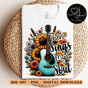 Pu&ograve; includere: Una maglietta bianca con una chitarra acustica blu e verde circondata da fiori colorati. Il testo "Then Sings My Soul" &egrave; scritto in lettere nere.