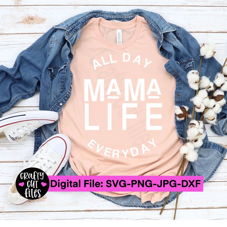 May include: A peach-colored t-shirt with the text "ALL DAY MAMA LIFE EVERYDAY" in white. The t-shirt is laid flat on a white wooden surface with a denim jacket, white sneakers, and cotton branches.