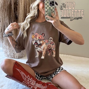 Puede incluir: Una camiseta marr&oacute;n con un estampado floral de un bulldog franc&eacute;s. La camiseta la lleva puesta una persona que lleva una gorra de b&eacute;isbol marr&oacute;n y blanca, botas de vaquero rojas y pantalones cortos de cuadros negros y blancos. El texto "luv juliette designs" es visible en la camiseta.