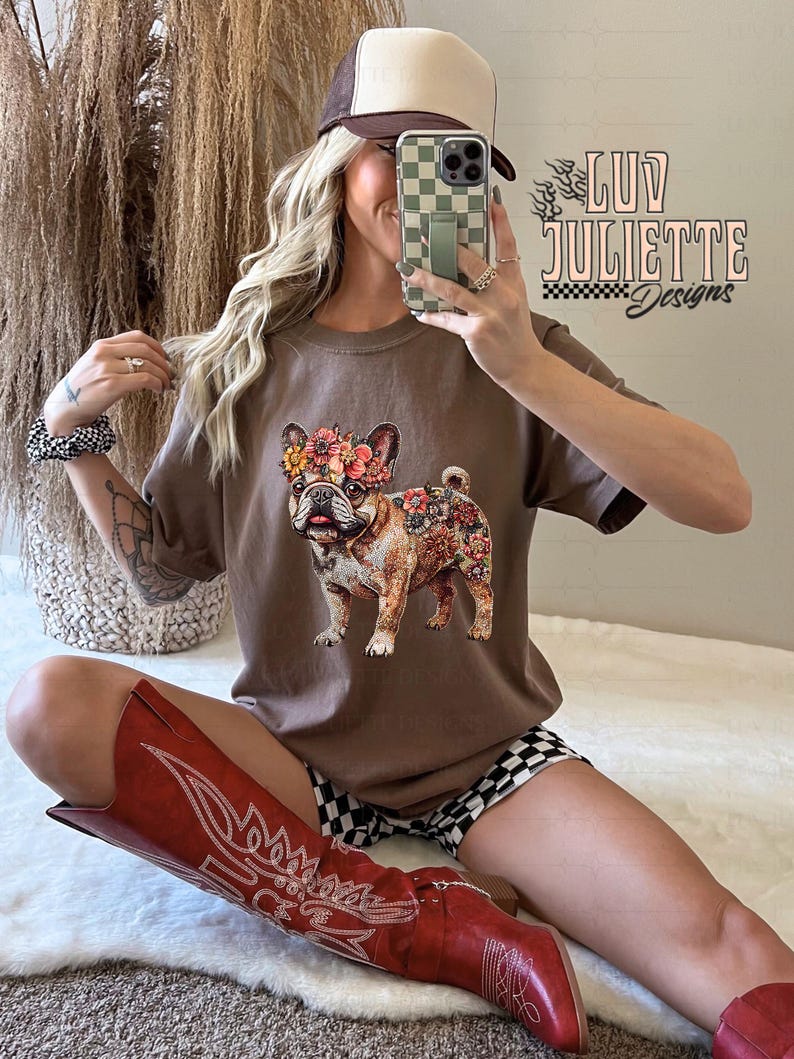 Puede incluir: Una camiseta marr&oacute;n con un estampado floral de un bulldog franc&eacute;s. La camiseta la lleva puesta una persona que lleva una gorra de b&eacute;isbol marr&oacute;n y blanca, botas de vaquero rojas y pantalones cortos de cuadros negros y blancos. El texto "luv juliette designs" es visible en la camiseta.