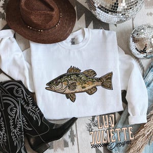 May include: White sweatshirt with a green and brown fish graphic. The sweatshirt has a crew neck and long sleeves. The fish is a bass with a detailed design. The text "Luv Juliette Designs" is printed on the bottom right corner of the sweatshirt.