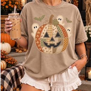 May include: A tan short-sleeve t-shirt featuring a large graphic of a patchwork-style jack-o'-lantern pumpkin. The pumpkin design includes various patterns like gingham, polka dots, and stripes, surrounded by small white ghosts and green bats.