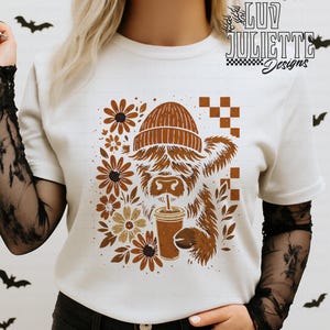 May include: White t-shirt with a brown Highland cow design wearing a beanie and drinking coffee. The design includes brown flowers and the text "Luv Juliette Designs". The shirt has long black lace sleeves.