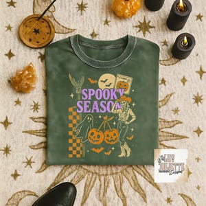 May include: Green crew neck sweatshirt with a Halloween-themed graphic. The design includes the words "Spooky Season", a ghost, pumpkins, bats, and a cowgirl. The sweatshirt has a vintage, washed look.