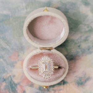 May include: An emerald-cut diamond ring with a halo of smaller stones set on a thin gold band, displayed inside an open, oval-shaped, cream-colored velvet ring box with a soft pink interior lining, showcasing a clear and elegant design.