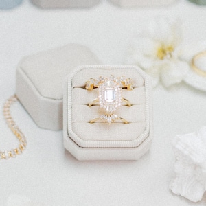 May include: An assortment of gold rings with diamond accents displayed in a white velvet ring box. The rings are surrounded by small, square, pastel-colored boxes. A gold chain and white flowers are also present, creating a delicate and elegant aesthetic.
