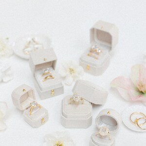 May include: An assortment of engagement rings and wedding bands displayed in open, light gray velvet ring boxes. The rings feature various diamond cuts and gold bands. White flowers and a small plate with rings are also present.