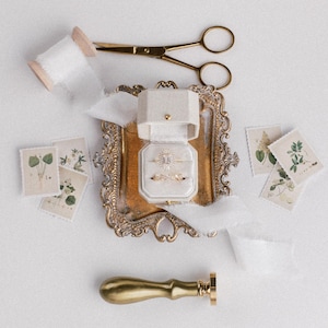 May include: An overhead shot of wedding-themed items on a white surface. A gold-colored ornate tray holds two rings in a ring box. Other items include a spool of white ribbon, gold scissors, botanical stamps, and a gold wax seal.