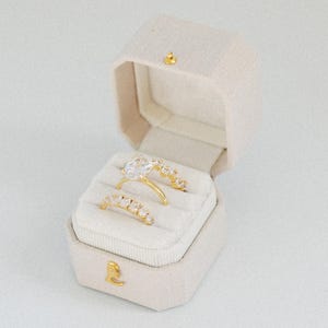 May include: A beige fabric ring box with a gold-tone latch containing two gold rings. The top ring features a large oval-cut clear stone with smaller stones along the band, while the bottom ring is a band set with multiple small clear stones.
