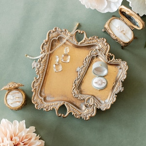 May include: A gold-toned, ornate tray holds jewelry, including a bracelet and square-cut earrings. Two ring boxes, one open, display rings with clear stones. The scene is set against a muted green backdrop, with soft pink and white flowers.