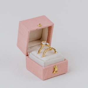 May include: A pink velvet ring box with a gold ring inside. The ring has a large, clear center stone and a band of smaller stones.