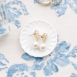 May include: An assortment of jewelry and accessories on a floral patterned surface. Includes a perfume bottle, a ring in a velvet box, two gold rings, and a small plate with pearl earrings. The background is white with blue floral patterns.