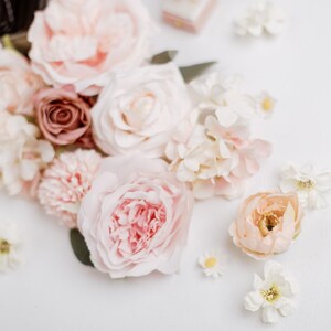 May include: An assortment of soft, pastel-colored flowers, including roses, peonies, and smaller blossoms, arranged on a white surface. The color palette includes shades of pink, peach, cream, and a touch of burgundy, creating a delicate and romantic aesthetic.