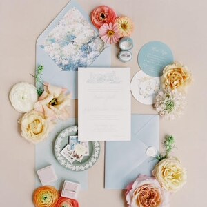 May include: A wedding stationery set featuring a light blue envelope with a floral liner, a white invitation card, a circular menu, and a small ring box. The arrangement is decorated with various fresh flowers including pink, yellow, and white roses.