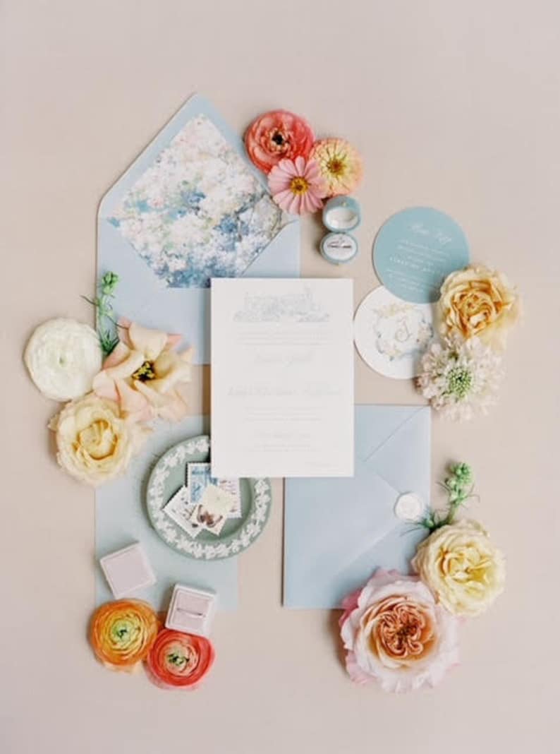May include: A wedding stationery set featuring a light blue envelope with a floral liner, a white invitation card, a circular menu, and a small ring box. The arrangement is decorated with various fresh flowers including pink, yellow, and white roses.