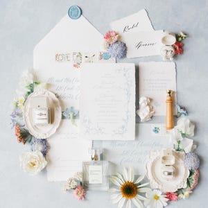 Puede incluir: Elegante conjunto de invitaciones de boda con detalles florales. Incluye tarjetas de invitaci&oacute;n, sobres, anillos en una caja, perfume y un sello de cera. La paleta de colores presenta azules suaves, blancos y rosas, con delicados arreglos florales.