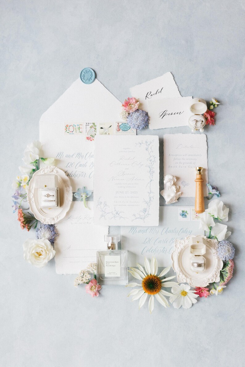 Puede incluir: Elegante conjunto de invitaciones de boda con detalles florales. Incluye tarjetas de invitaci&oacute;n, sobres, anillos en una caja, perfume y un sello de cera. La paleta de colores presenta azules suaves, blancos y rosas, con delicados arreglos florales.