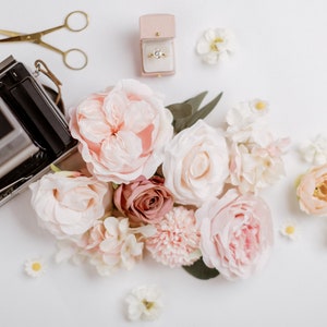 May include: An arrangement of soft pink and white flowers, a vintage camera, gold scissors, and a ring in a pink box on a white surface. The flowers include roses and smaller blossoms, creating a romantic and delicate aesthetic.