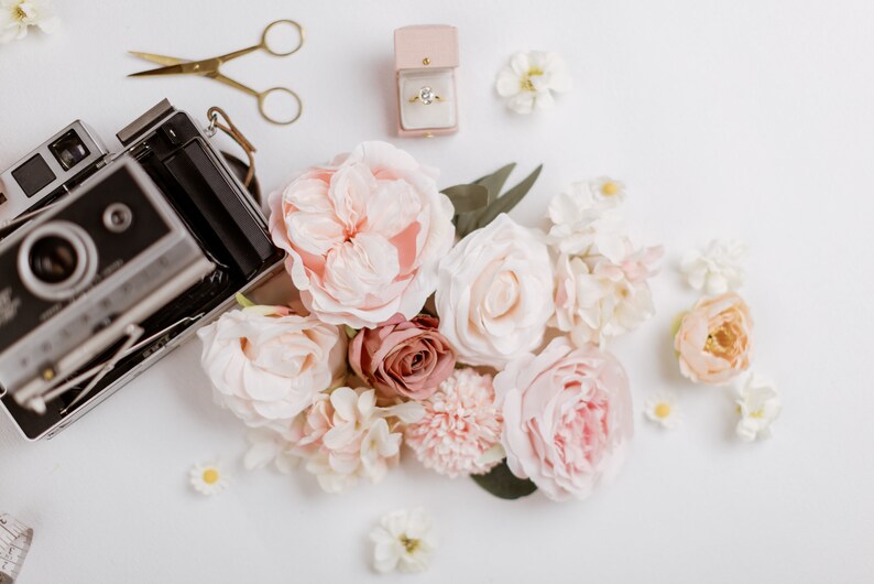 May include: An arrangement of soft pink and white flowers, a vintage camera, gold scissors, and a ring in a pink box on a white surface. The flowers include roses and smaller blossoms, creating a romantic and delicate aesthetic.