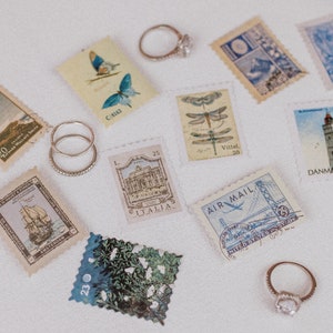 May include: An assortment of vintage postage stamps with various designs, including butterflies, dragonflies, and architectural landmarks. Several rings with clear gemstones and a band of small stones are also present, arranged on a white surface.