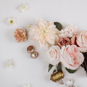 May include: A flat lay featuring a collection of blush and cream-colored flowers, including roses and dahlias, alongside a vintage camera, a ring box with a diamond ring, and a decorative gold bell. Scattered white flowers complete the composition.
