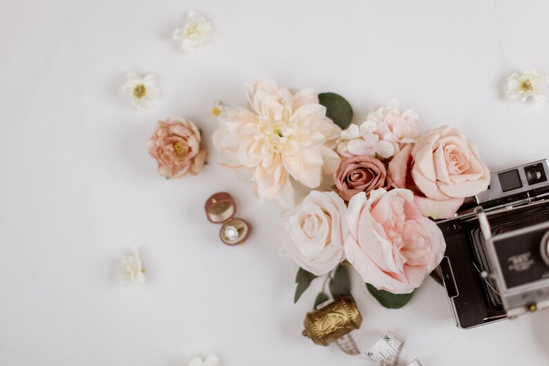 May include: A flat lay featuring a collection of blush and cream-colored flowers, including roses and dahlias, alongside a vintage camera, a ring box with a diamond ring, and a decorative gold bell. Scattered white flowers complete the composition.