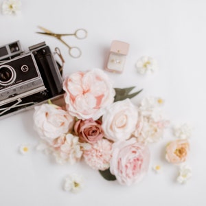 May include: A vintage Polaroid camera sits among a collection of pastel-colored roses and other flowers. A small pair of gold scissors and a ring box with a ring are also present. The scene is arranged on a white surface, creating a soft, romantic aesthetic.