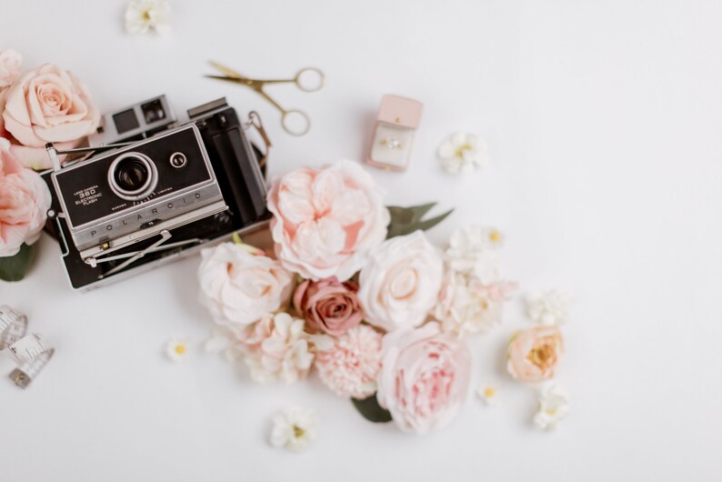 May include: A vintage Polaroid camera sits among a collection of pastel-colored roses and other flowers. A small pair of gold scissors and a ring box with a ring are also present. The scene is arranged on a white surface, creating a soft, romantic aesthetic.