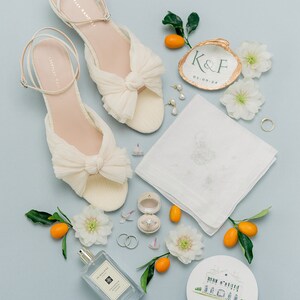 May include: A pair of cream-colored pleated sandals with ankle straps, a white embroidered handkerchief, a perfume bottle, a ring in a velvet box, two wedding bands, a painted shell with K&F 03-09-24, a round card with a house illustration, and kumquats.